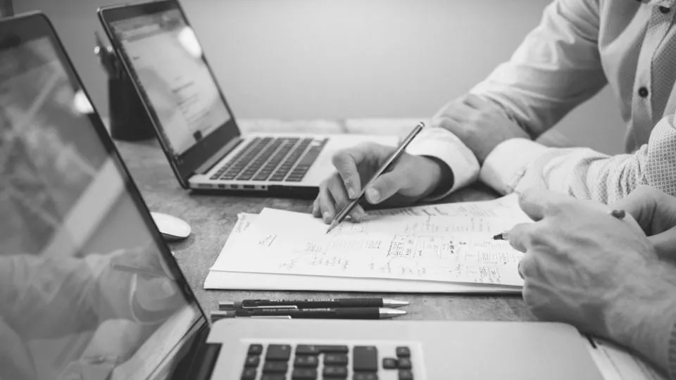 Duas pessoas à mesa, vistas só pelas mãos, fazem anotações em folhas de papel entre dois computadores portáteis