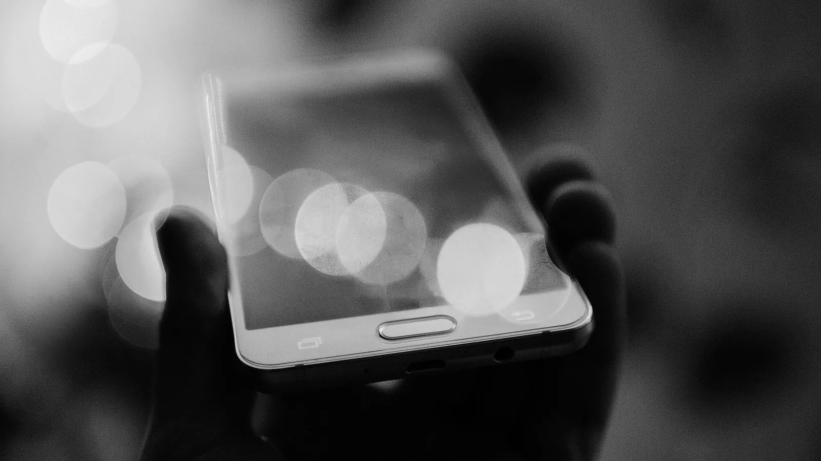 Mão segurando um celular com luzes desfocadas refletidas na tela, sem que se veja o que ela mostra