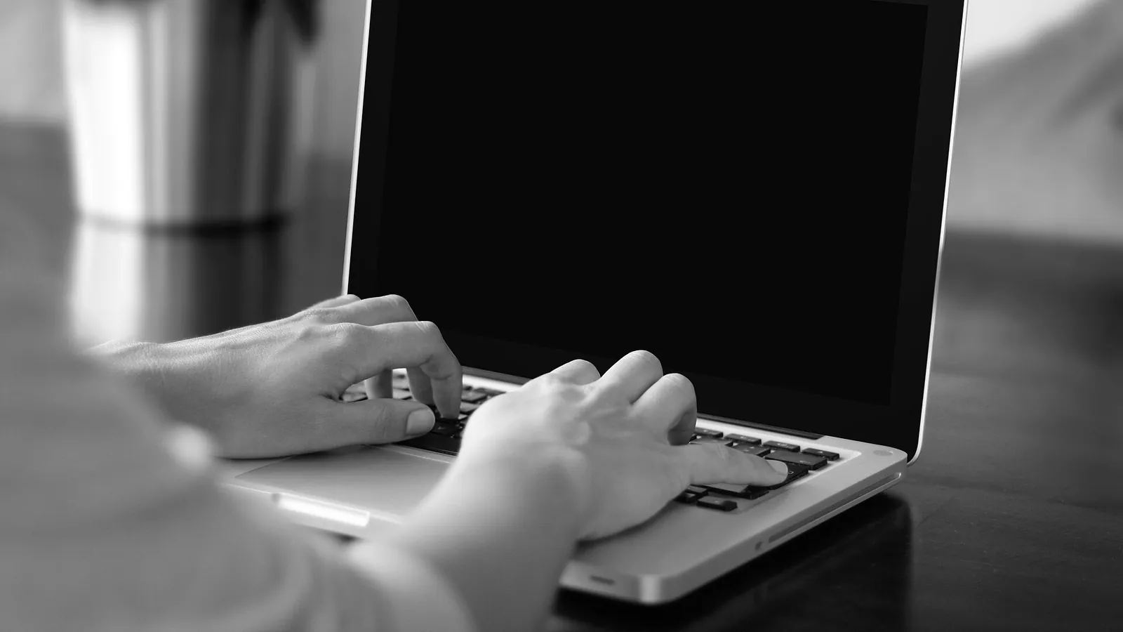 Mãos digitando num notebook de tela escura, apoiado numa mesa de madeira
