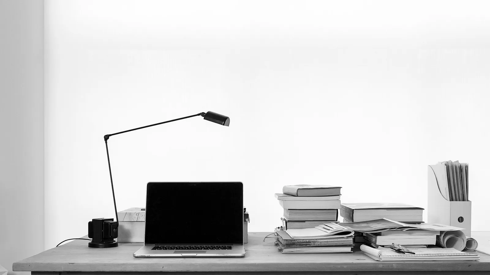 Mesa de trabalho com notebook, luminária e pilhas de livros e papéis, sem ninguém sentado