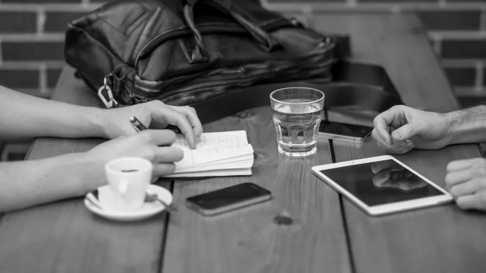 Mãos de duas pessoas em uma mesa de café, com celulares, um tablet e um caderno de anotações