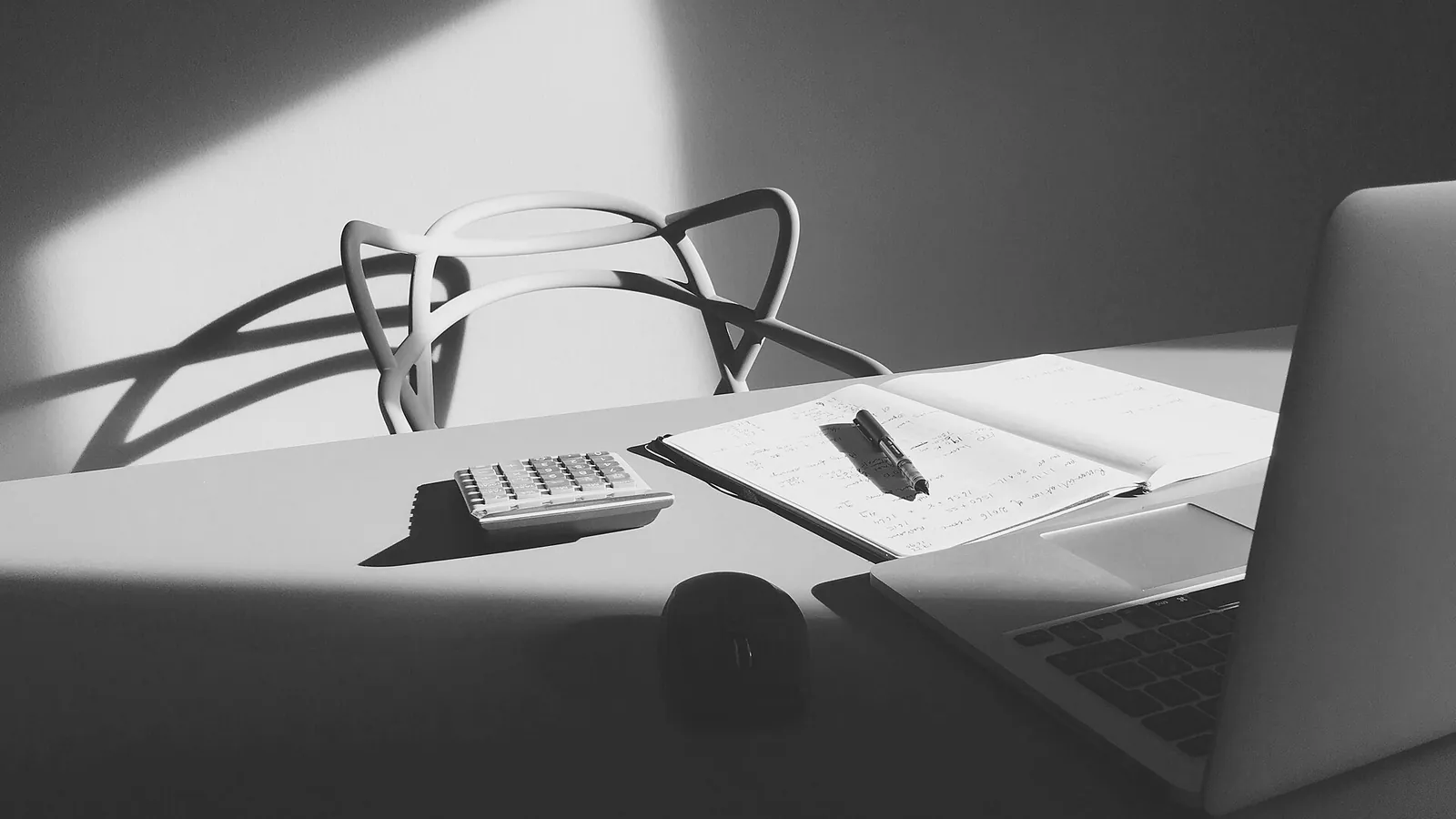 Mesa de escritório com caderno de contas aberto, caneta, calculadora e computador portátil, diante de uma cadeira vazia