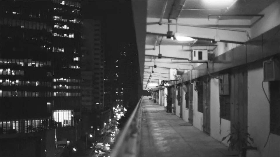 Corredor externo de um prédio de apartamentos com as luzes acesas, as portas de um lado e a cidade escura do outro