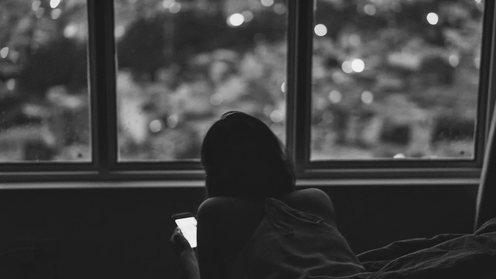 Pessoa vista de costas, deitada diante de uma janela à noite, olhando a tela acesa do celular