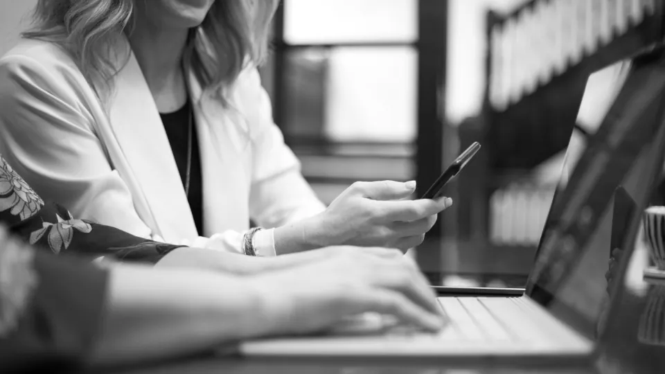 Mulher de blazer claro segurando o celular diante de um computador portátil, em uma mesa de escritório, com o rosto fora do quadro