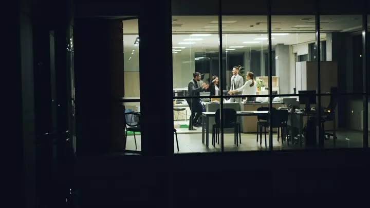 Escritório iluminado à noite visto de fora, com pessoas trabalhando